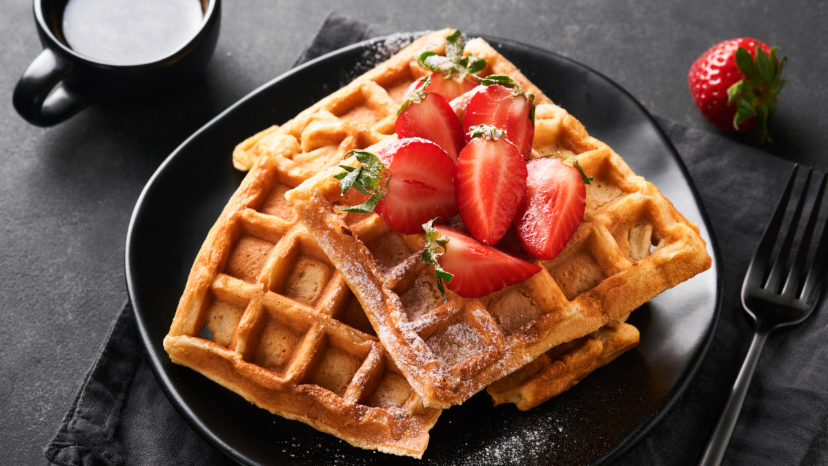 Belgium waffles. Homemade waffles with strawberries, powdered sugar and cup of coffee on black plate on black stone table background. Breakfast. Top view. Mockup for design idea.