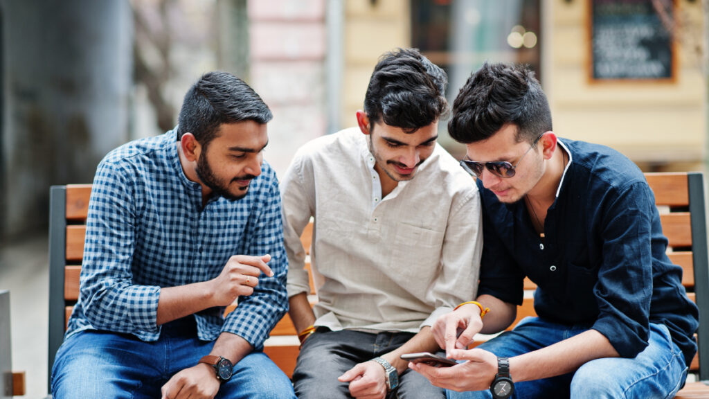 Group of three indian ethnicity friendship togetherness mans. Technology and leisure, guys with phones, sitting on bench.